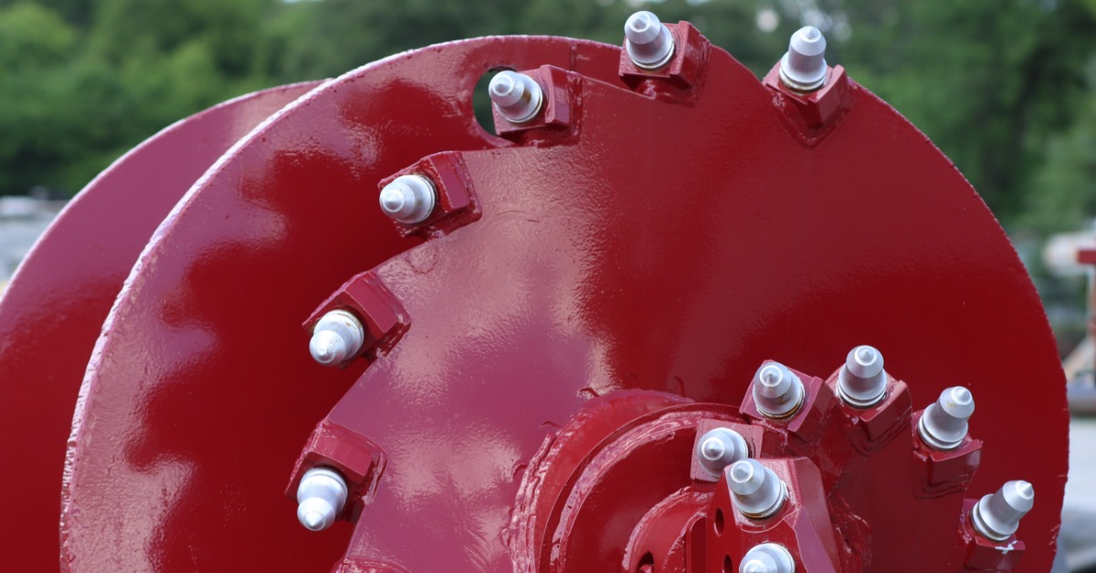 A red metal auger with silver cutting teeth arranged along curved blades against a blurred green outdoor background.